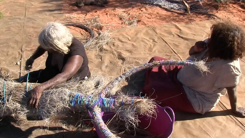 'Using blue wool for added colour, Angilyiya ties the <em>tjanpi</em> (grass) together. She leads the group in singing the Kuru Ala <em>inma</em> (ceremonial song).'