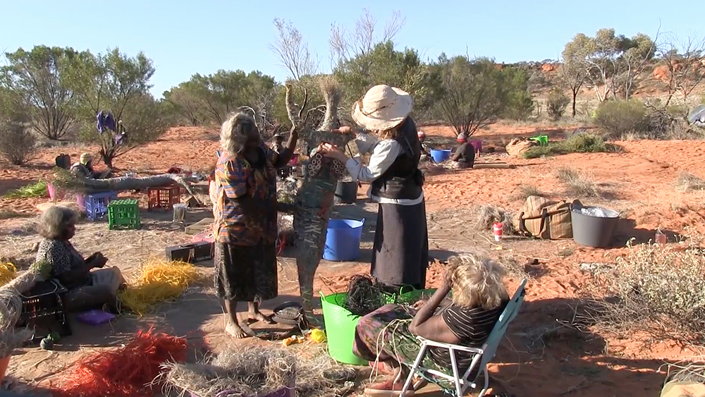 'Anawari and Jo Foster, of Tjanpi Desert Weavers, assess progress and the orientation of the <em>yipi</em> (breasts). The artists decide how to outline the <em>yipi</em> using a coil of yellow raffia and <em>tjanpi</em> (grass).'