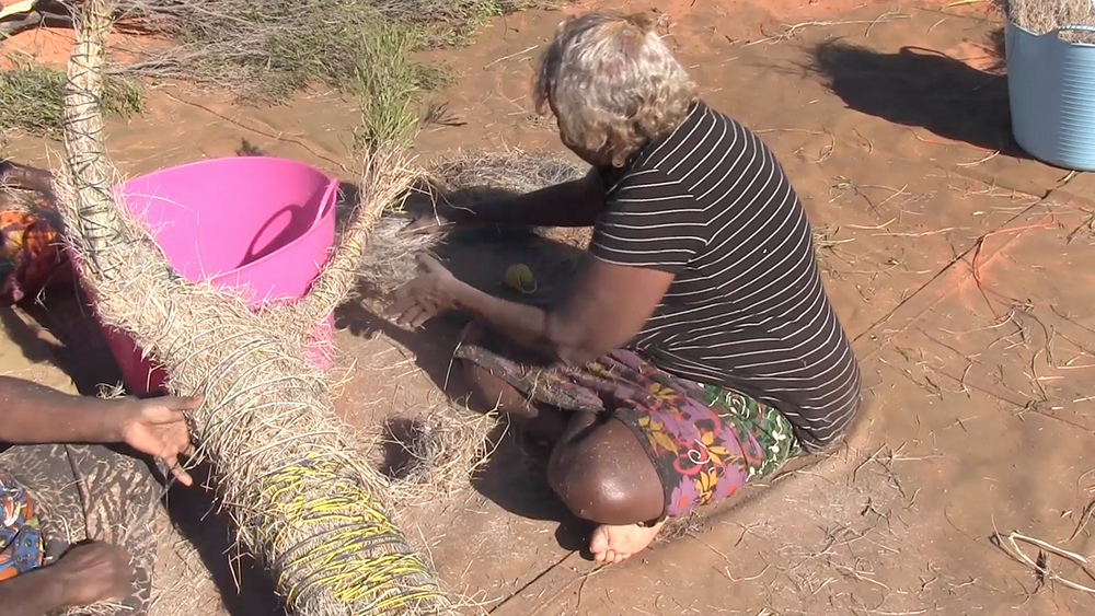 'Anawari uses wool to attach a layer of grass around the body of the figure and Nora rubs the grass to prepare it. Gradually the sister takes shape as they secure the handfuls of grass by tightly winding wool and raffia over and around the figure.'