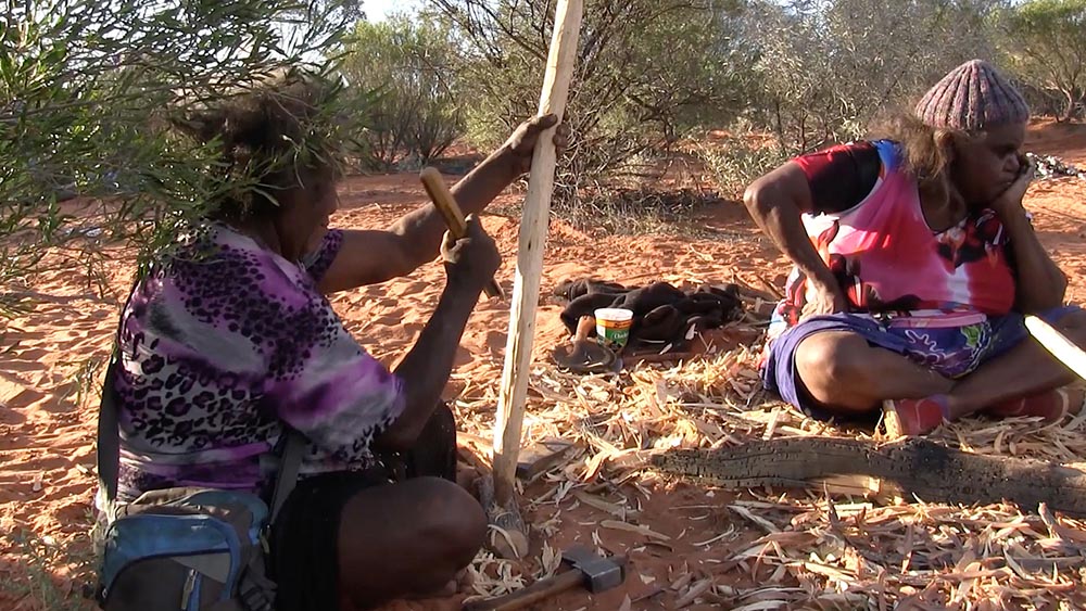 'Janet Nyumitji Forbes chips away at a piece of wood to make a <em>wana</em>, used by desert women to collect bush foods such as <em>maku</em> (witchetty grubs) and <em>mirrka</em> (plant foods). Once the <em>wana</em> has been formed, Yayimpi Lewis smooths the surface using a glass shard.'